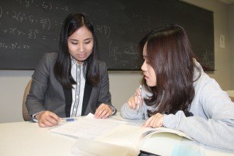 Two asian female students work together, one tutoring the other with the aid of reference books and notes, the board behind them filled with formulas. You too can engage in learning with others at ܽƵ Michigan University. 
